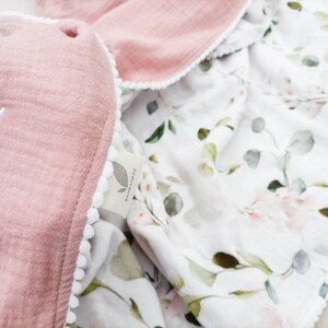 May include: A pink muslin baby blanket with white pom pom trim and a floral print on the reverse side. The blanket is personalized with the name "Ada" embroidered on the pink side.