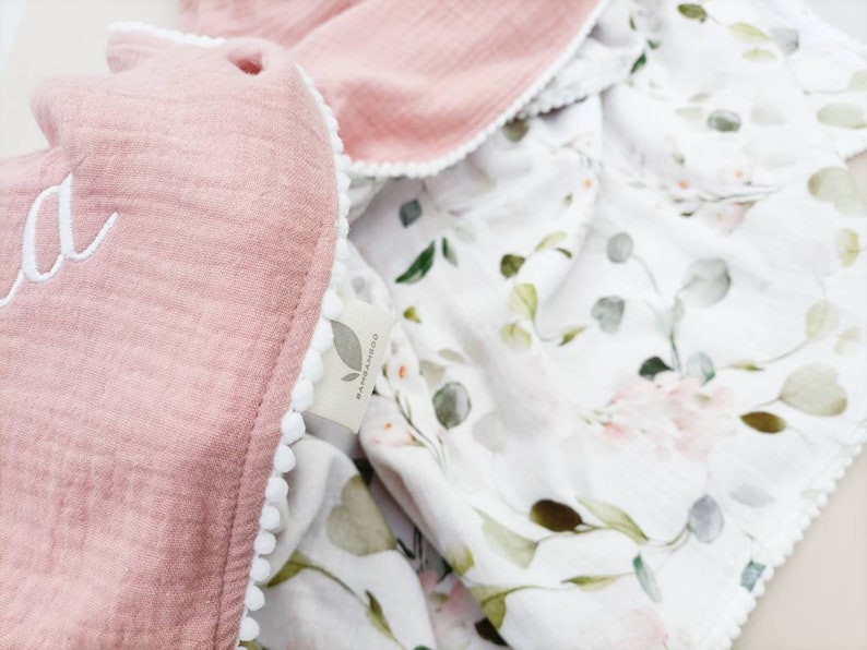 May include: A pink muslin baby blanket with white pom pom trim and a floral print on the reverse side. The blanket is personalized with the name "Ada" embroidered on the pink side.