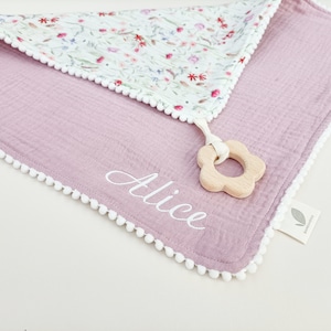 Op de afbeelding: Een lavendelkleurige babydeken met een bloemenpatroon op de bovenlaag en witte pompomrand. De naam "Alice" is in witte letters geborduurd. Een houten bloemvormige bijtring is bevestigd. Het label luidt "Bambamdoo".
