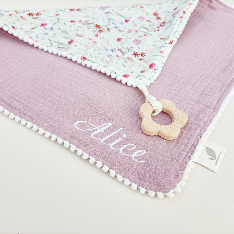 Op de afbeelding: Een lavendelkleurige babydeken met een bloemenpatroon op de bovenlaag en witte pompomrand. De naam "Alice" is in witte letters geborduurd. Een houten bloemvormige bijtring is bevestigd. Het label luidt "Bambamdoo".