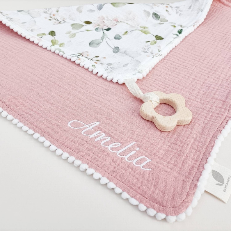 Op de afbeelding: Een roze babydeken met een bloemenpatroon op de bovenlaag en witte pompom-afwerking. De naam "Amelia" is in het wit op de deken geborduurd. Een houten bloemvormige bijtring is bevestigd.