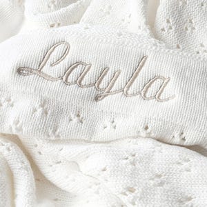 May include: White crocheted baby blanket with the name "Layla" embroidered in a light brown thread.