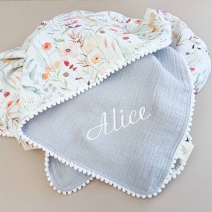 May include: A baby blanket with a floral pattern and a gray interior. The blanket features a white pom-pom trim and the name "Alice" embroidered in white script. The floral pattern includes orange, red, and purple flowers.