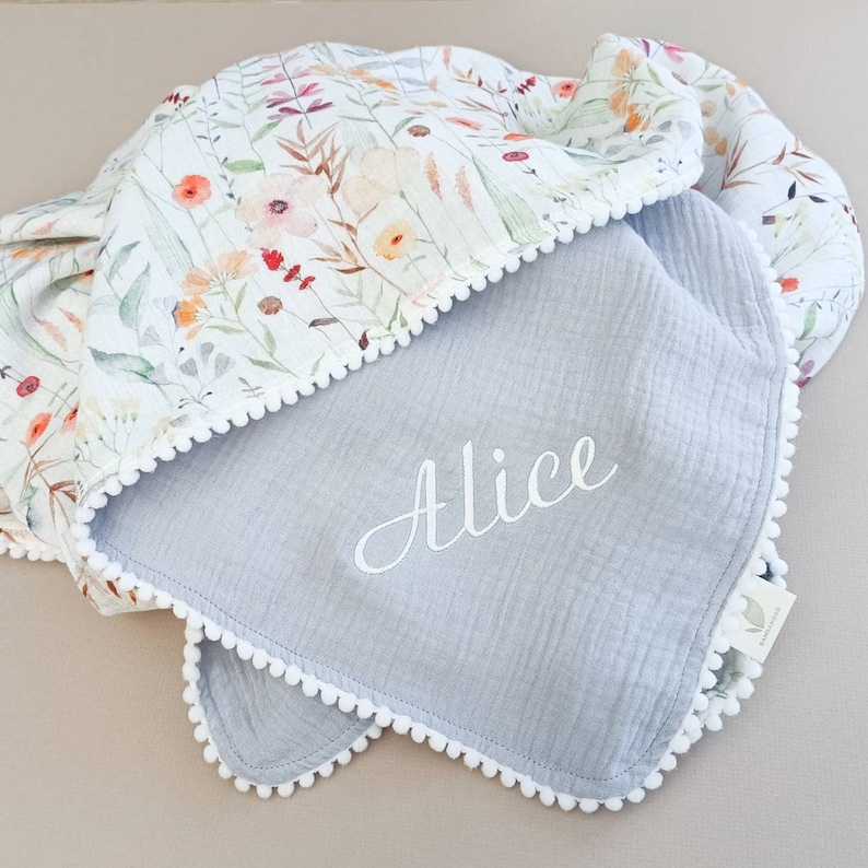 May include: A baby blanket with a floral pattern and a gray interior. The blanket features a white pom-pom trim and the name "Alice" embroidered in white script. The floral pattern includes orange, red, and purple flowers.
