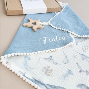 Op de afbeelding: Een babydeken met een blauwe rand en witte pompom-afwerking, met een oceaan-thema print. Een houten zeester-bijtring is bevestigd. De naam "Finley" is op de deken geborduurd. Een kartonnen doos met de merknaam "Bambamboo" staat op de achtergrond.