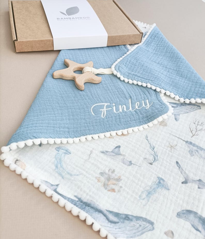 Op de afbeelding: Een babydeken met een blauwe rand en witte pompom-afwerking, met een oceaan-thema print. Een houten zeester-bijtring is bevestigd. De naam "Finley" is op de deken geborduurd. Een kartonnen doos met de merknaam "Bambamboo" staat op de achtergrond.