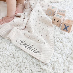 May include: A cream-colored knitted baby blanket with a delicate openwork pattern. The blanket is personalized with the name "Arthur" embroidered in black thread. The blanket is lying on a white fluffy rug with wooden blocks spelling out the name "Max" in blue and brown letters.