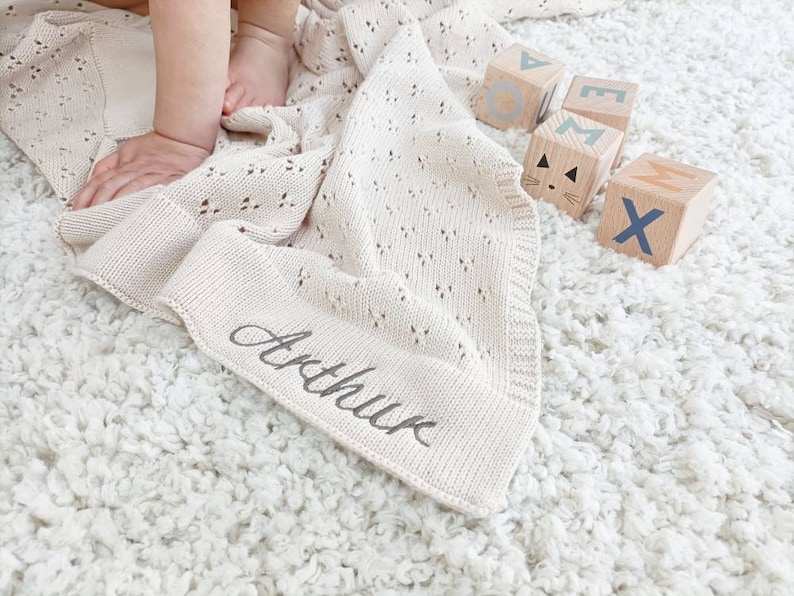 May include: A cream-colored knitted baby blanket with a delicate openwork pattern. The blanket is personalized with the name "Arthur" embroidered in black thread. The blanket is lying on a white fluffy rug with wooden blocks spelling out the name "Max" in blue and brown letters.