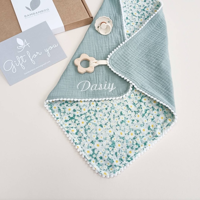 Op de afbeelding: Een baby slabbetje met een bloemenpatroon en een effen teal achterkant. De slab heeft een witte pompom-rand en een houten bloemvormige bijtring. Een fopspeen en een cadeaukaart zijn ook zichtbaar. De naam "Dasiy" is op de slab geborduurd.
