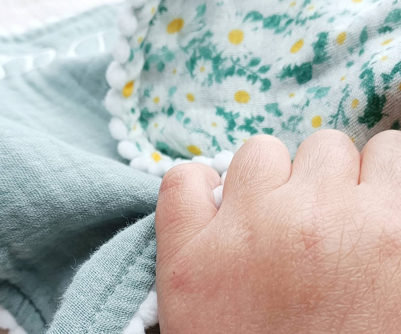 Op de afbeelding: Close-up van een zachte, lichtgroene stof met een getextureerde uitstraling. Een gedeelte van stof met een bloemenpatroon in wit, geel en groen is zichtbaar. Witte pomponranden omzomen de bloemenstof. De afbeelding toont een hand die de stof vasthoudt.