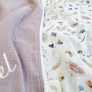 May include: A soft, lavender-colored blanket with the name "Isabel" embroidered in white script. The blanket's edge is lined with a white pom-pom trim. The reverse side features a floral pattern with butterflies and botanical elements.