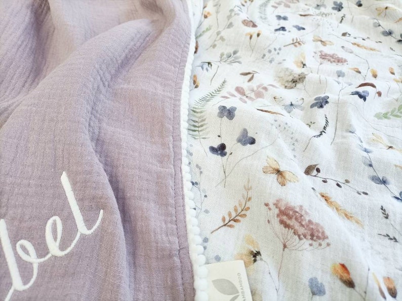 May include: A soft, lavender-colored blanket with the name "Isabel" embroidered in white script. The blanket's edge is lined with a white pom-pom trim. The reverse side features a floral pattern with butterflies and botanical elements.