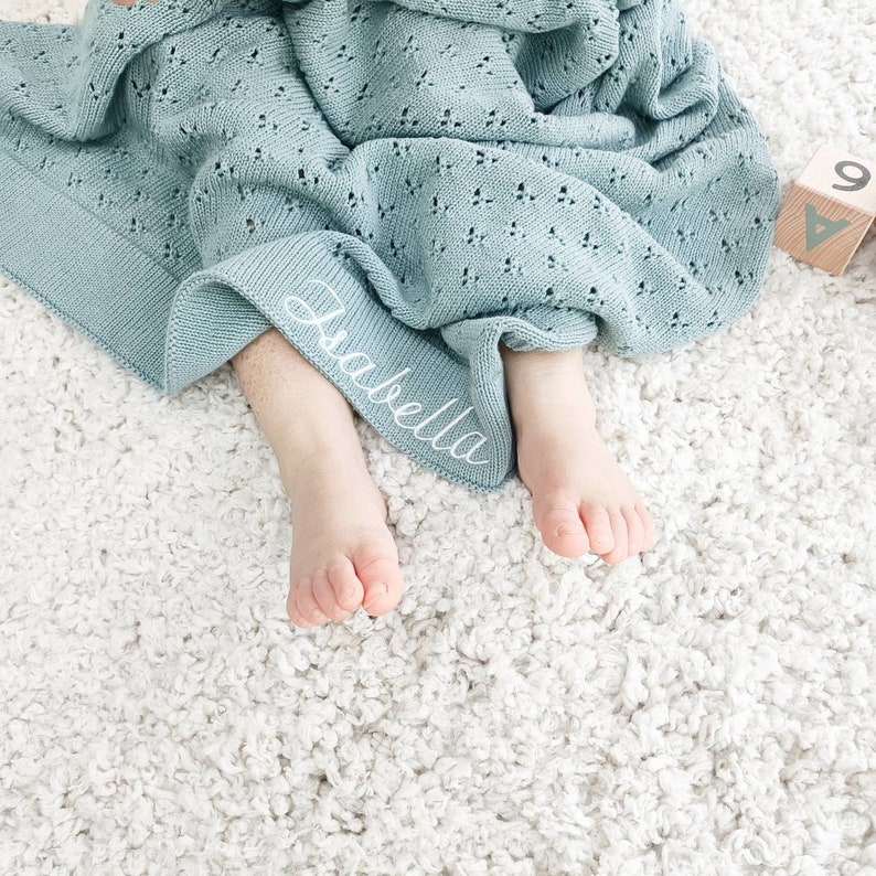 May include: A light teal knitted baby blanket with a delicate openwork pattern. The blanket has a personalized embroidered name, "Isabella", in white script. The blanket is draped over a baby's feet on a white, textured surface.