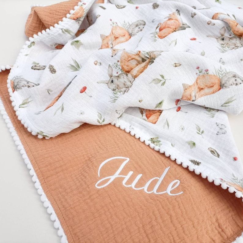May include: A baby blanket with a woodland animal print featuring foxes, rabbits, and hedgehogs on a white background. The blanket has a rust-colored border with white pom-pom trim and the name "Jude" embroidered in white.