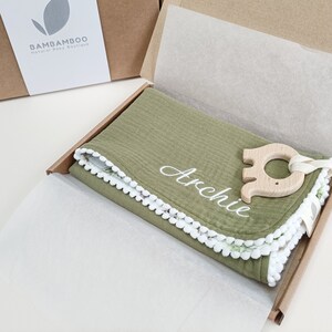 Op de afbeelding: Een babycadeauset in een kartonnen doos. De set bevat een groene deken met witte geborduurde tekst "Archie", een houten olifant bijtring en een witte pompom afwerking. De doos bevat ook wit vloeipapier.