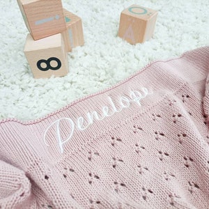 May include: A pink knitted baby blanket with a white embroidered name "Penelope" on it.