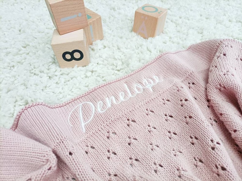May include: A pink knitted baby blanket with a white embroidered name "Penelope" on it.