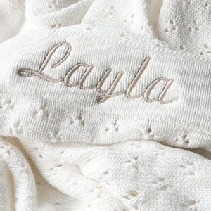 May include: White crocheted baby blanket with the name "Layla" embroidered in beige thread.
