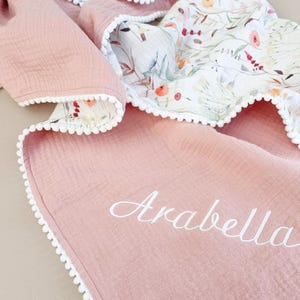 May include: A pink baby blanket with a floral pattern on one side and the name "Arabella" embroidered in white. The blanket has a white pom-pom trim.