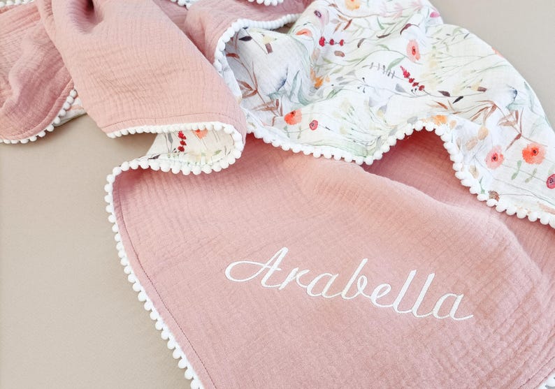 May include: A pink baby blanket with a floral pattern on one side and the name "Arabella" embroidered in white. The blanket has a white pom-pom trim.