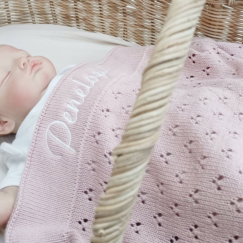 May include: A pink knitted baby blanket with a white embroidered name "Perdita" on it. The blanket is in a wicker basket.