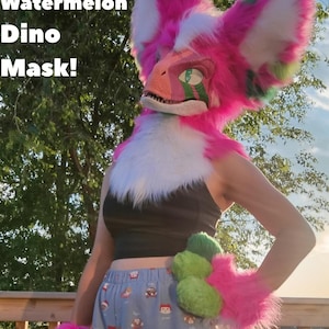 May include: A Watermelon Dino Mask with bright pink and white faux fur, featuring a reptilian face with green accents. The set includes matching pink and green furry hand paws. The text Watermelon Dino Mask! and Professionally Made is visible.