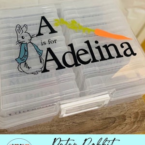 May include: A clear plastic photo organizer with a Peter Rabbit design. The lid has a personalized label with the letter A, the words "is for" and the name "Adelina".
