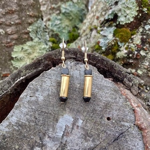 May include: A pair of gold earrings with black bullet-shaped charms. The earrings are hanging from a wooden surface.