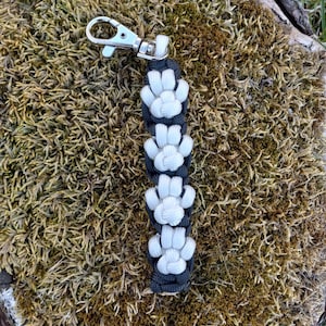 May include: A black and white paracord keychain with a silver key ring. The keychain is made of a series of white paw prints.