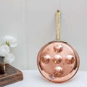 Puede incluir: Una sartén de cobre para caracoles con cinco hendiduras y un asa de latón. La sartén redonda se muestra sobre una superficie blanca. El fondo incluye una pared blanca y una tabla de cortar de madera.