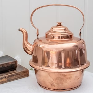 Puede incluir: Una tetera de cobre con un pico curvo, una tapa abovedada y un asa grande. La tetera está sobre una superficie blanca junto a dos libros antiguos. La tetera es de color cobre brillante y reflectante.