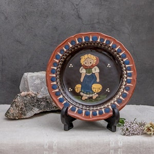 May include: A brown ceramic plate with a blue and red border. The plate features a hand-painted design of a girl in a blue dress carrying two yellow buckets.