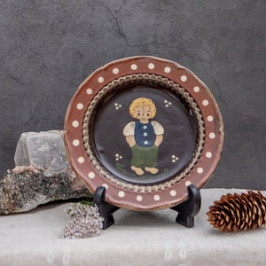 Op de afbeelding: Een bruin keramiek bord met een witte rand en witte stippen. Het bord heeft een handgeschilderde illustratie van een kind dat een blauw shirt en een groene broek draagt. Het bord staat op een standaard en wordt gepresenteerd op een wit linnen tafelkleed.