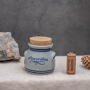 Op de afbeelding: Een kleine blauwe en witte keramische pot met een kurken deksel. De pot is gelabeld met "Paprika" en heeft een blauw en wit ontwerp. Een kleine kurk met de tekst "Beaune France" staat naast de pot.
