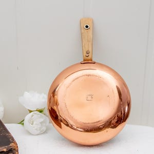 Puede incluir: Una sartén de cobre con mango de madera. La sartén tiene una superficie brillante y reflectante y un mango de madera clara sujeto con dos remaches. El mango mide aproximadamente 15 cm de largo. La sartén se utiliza probablemente para cocinar.