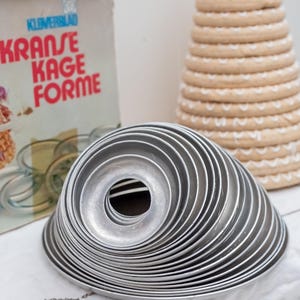 Könnte beinhalten: Ein Satz silberner Kransekake-Formen aus Metall, gestapelt auf einem weißen Tuch. Im Hintergrund befindet sich ein kegelförmiger Stapel Backwaren, der mit einer kleinen norwegischen Flagge gekrönt ist. Eine Box mit dem Text "KRANSE KAGE FORME" ist ebenfalls sichtbar.