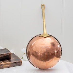 May include: A copper saucepan with a brass handle. The pan has a rounded bowl shape and a long, slender handle with a loop at the end. The copper surface reflects light, and the handle is a contrasting gold color.