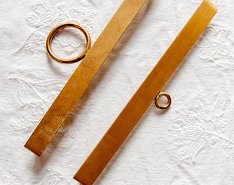 May include: Two gold-colored, rectangular drawer pulls with small, round handles. The pulls are made of a warm-toned metal and are set against a white, textured background. The design is simple and elegant, suitable for various furniture styles.