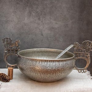 Op de afbeelding: Een zilverkleurige soepkom met twee sierlijke handvatten, elk met een ontwerp van een paardenkoets. Een bijpassende lepel rust in de kom. De kom heeft een getextureerd oppervlak en staat op een witte doek.