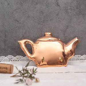 Puede incluir: Un porta llaves con forma de tetera de color cobre con tres ganchos. El porta llaves está sobre una superficie blanca con un borde de encaje.