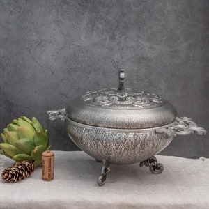 Può includere: Un piatto da portata in argento con un coperchio e un design decorato. Il piatto ha tre piedi e due manici. Il piatto è appoggiato su una tovaglia bianca con un carciofo verde e un tappo di vino.