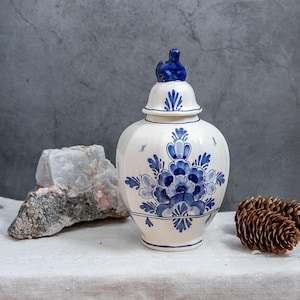 Può includere: Un vaso di ceramica bianco con coperchio, ornato da motivi floreali blu. Il vaso ha una forma bombata e una piccola maniglia blu sul coperchio. Il design ricorda la ceramica di Delft.
