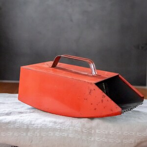 May include: A red metal scoop with a handle. The scoop is slightly curved and has a black interior. The scoop is sitting on a white fabric surface.
