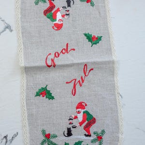 Könnte beinhalten: Grauer Tischläufer aus Leinen mit gestickten Weihnachtsmotiven, darunter Weihnachtsmänner, Stechpalmen und die roten Worte "God Jul". Der Läufer hat eine cremefarbene Spitzenborte, die eine dekorative Note verleiht.