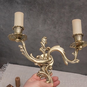Könnte beinhalten: Eine goldfarbene Wandleuchte aus Metall mit zwei Kerzenhaltern. Die Leuchte hat ein dekoratives, verziertes Design mit geschwungenen Mustern.
