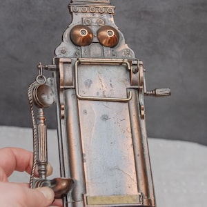 Op de afbeelding: Antiek stijl wandtelefoon met een messing afwerking. De telefoon heeft een draaischijf en een hoorn met een snoer. De telefoon is gemonteerd aan een muur met een decoratief scrollontwerp.