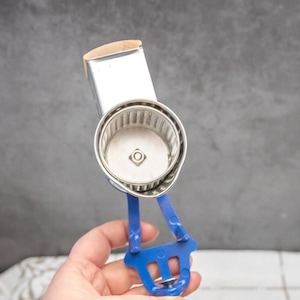 May include: A vintage food mill with a stainless steel grinding chamber and a blue plastic handle. The mill has a wooden top and a silver metal handle. This kitchen tool is designed for pureeing fruits and vegetables.