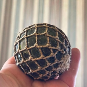Peut inclure: Une sphère en verre vert foncé enveloppée dans un filet décoratif en corde de jute beige. La surface du verre semble lisse et arrondie, tandis que la ficelle nouée crée un motif en grille autour de l'extérieur de l'orbe, avec des fibres effilochées.