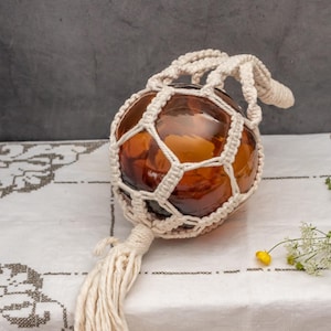 Puede incluir: Una bola decorativa de vidrio ámbar está encerrada en una red de macramé color crema con una borla larga. La bola de vidrio tiene forma redonda y está suspendida en el centro del macramé. El fondo es un mantel blanco.