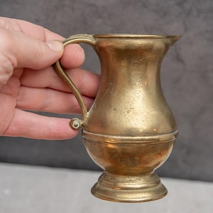Puede incluir: Jarra pequeña de latón antiguo con asa y base redondeada. La jarra tiene un borde ligeramente ensanchado y un asa decorativa. El latón tiene un color dorado cálido y muestra signos de edad y desgaste. La jarra está sujeta por una mano.